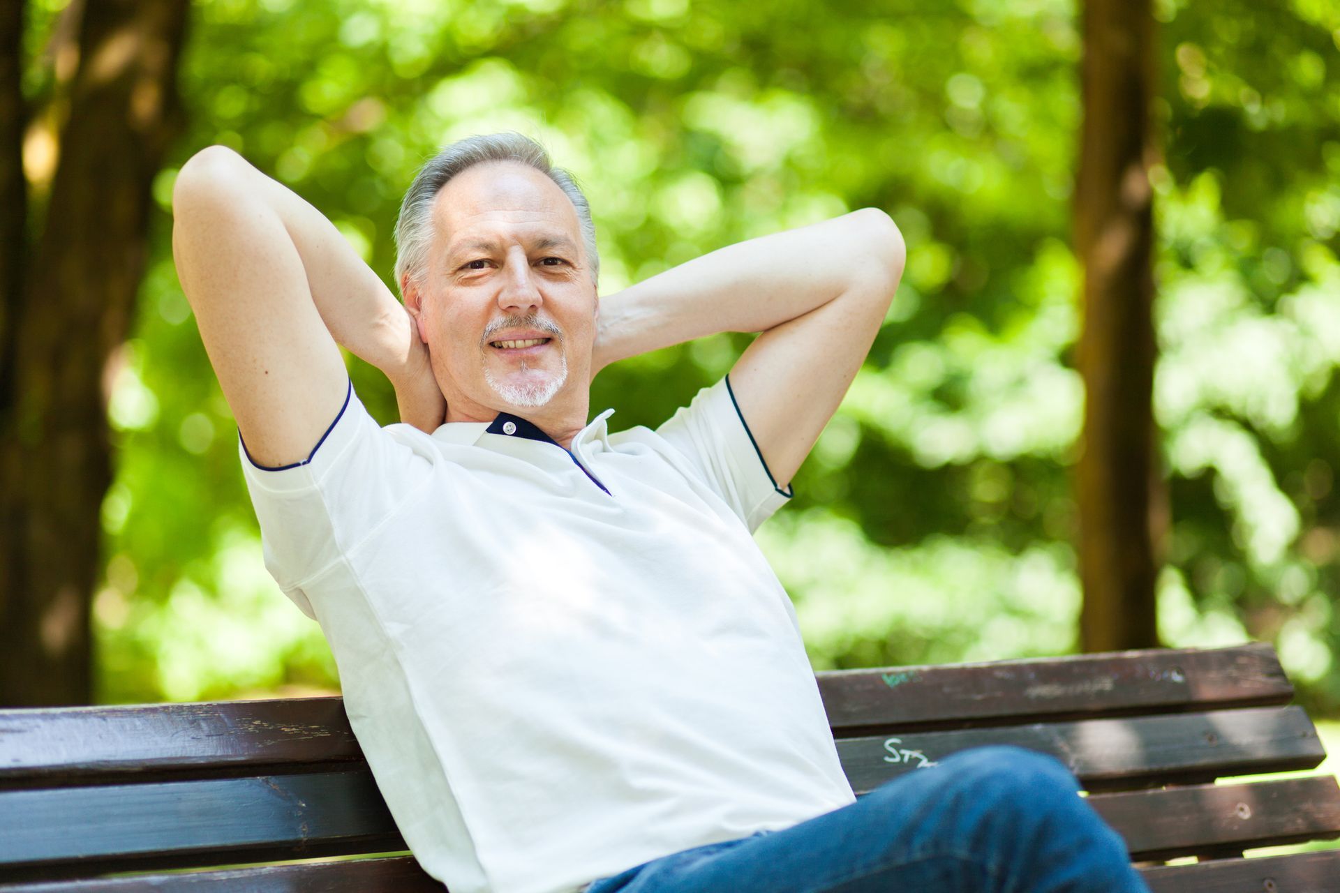 Man with gray hair and a white shirt relaxes on a park bench with hands behind his head. Man with gray hair and a white shirt relaxes on a park bench with hands behind his head.