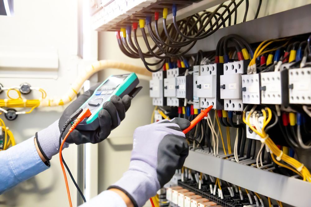Electrician testing wires in a control panel with a multimeter, wearing gloves.