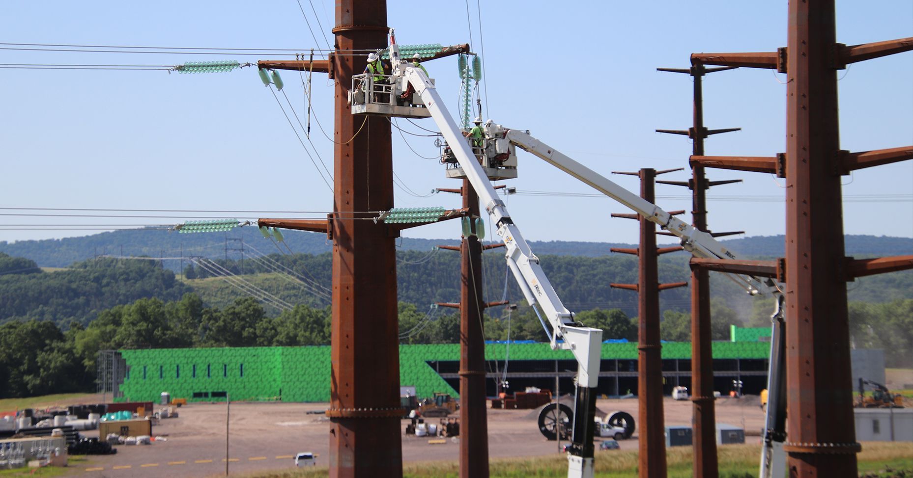 Workers in a lift servicing high-voltage power lines on a steel tower; bright sky and trees in background.