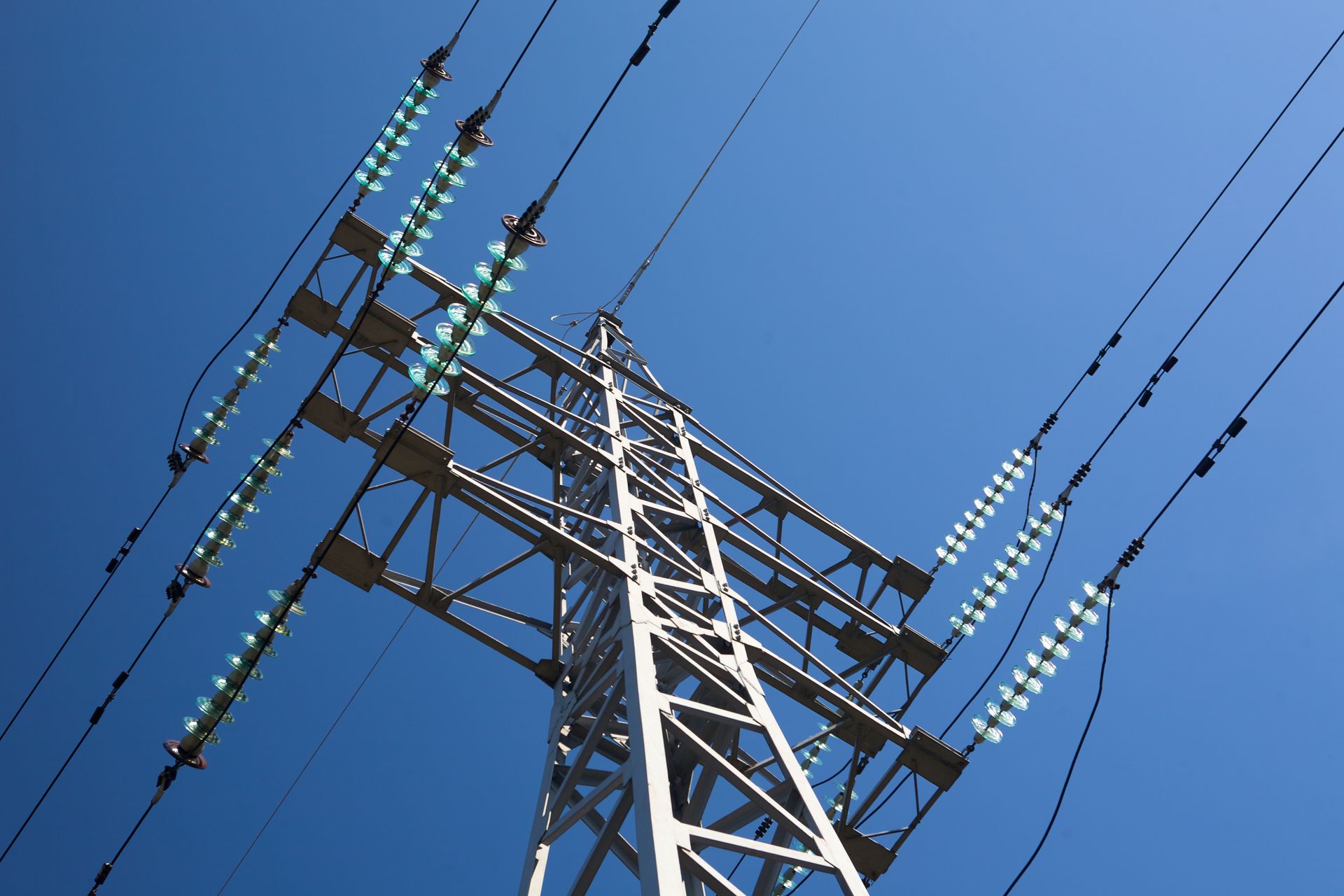 Workers in a lift servicing high-voltage power lines on a steel tower; bright sky and trees in background.