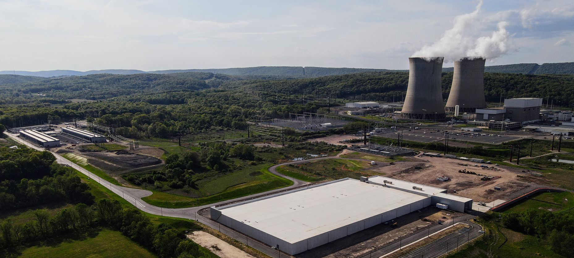 Aerial view of a nuclear power plant with two cooling towers releasing steam. Lush green forest surrounds the facility.