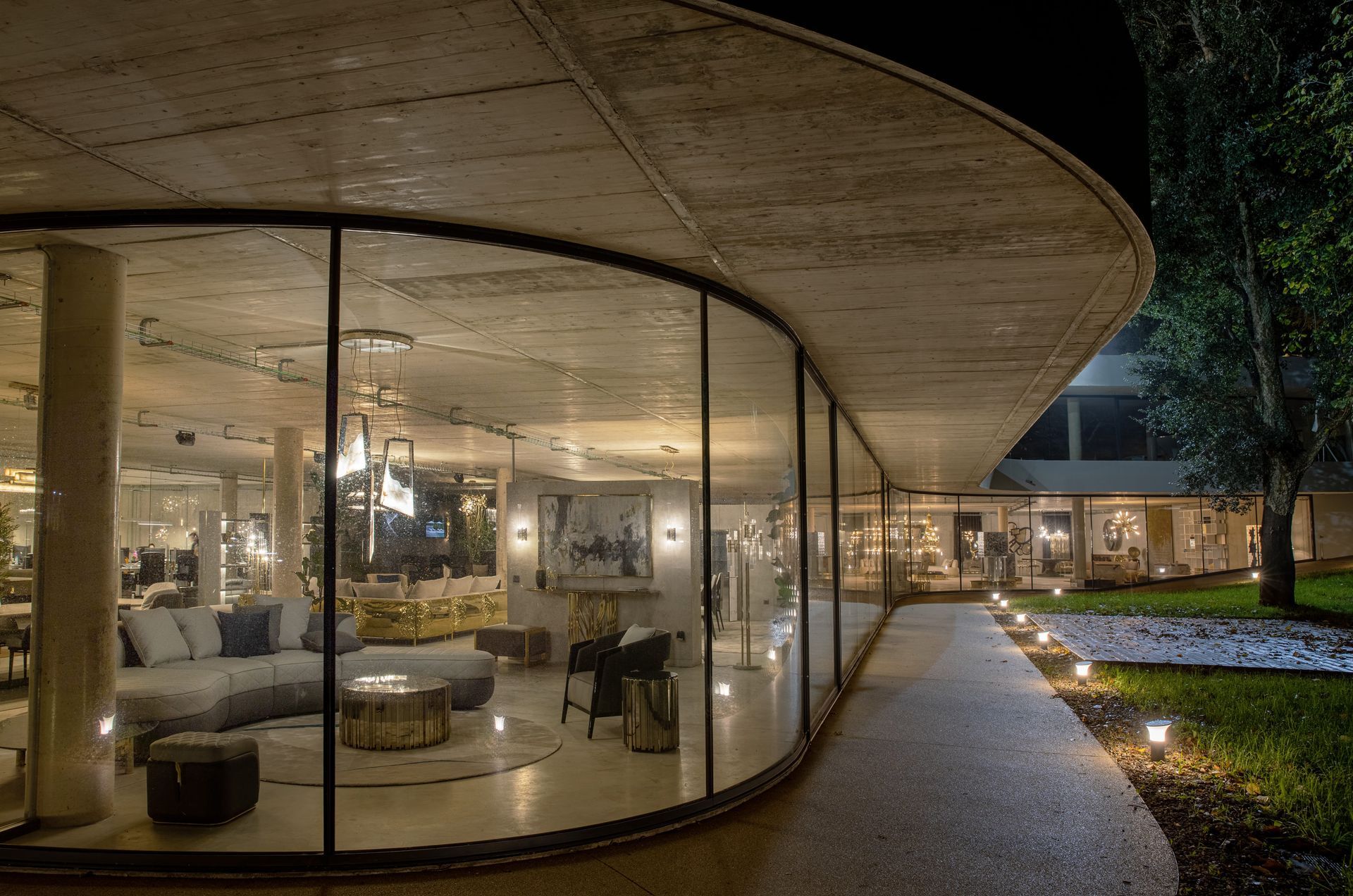 An elegant building with a curved glass facade by OTIIMA, warmly lit interior, and an inviting pathway illuminated by soft garden lights at night.