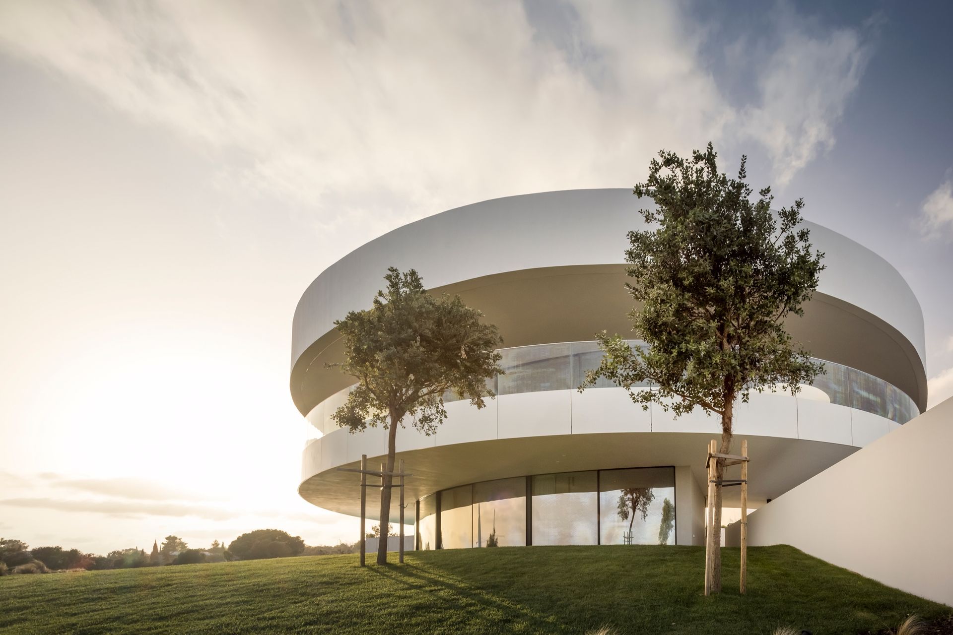 A contemporary circular building featuring sleek, curved windows by OTIIMA, set against a backdrop of a cloudy sky and surrounded by well-manicured lawns and trees.