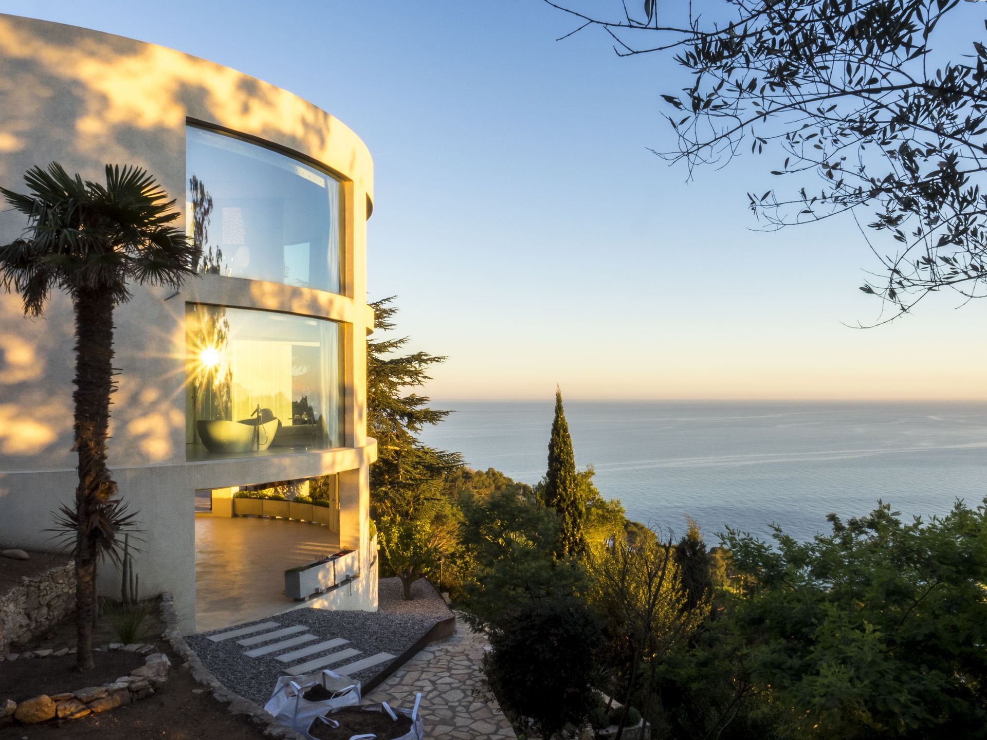 A modern house with expansive OTIIMA curved windows overlooking a serene coastal view, framed by lush greenery and a clear blue sky at sunset.