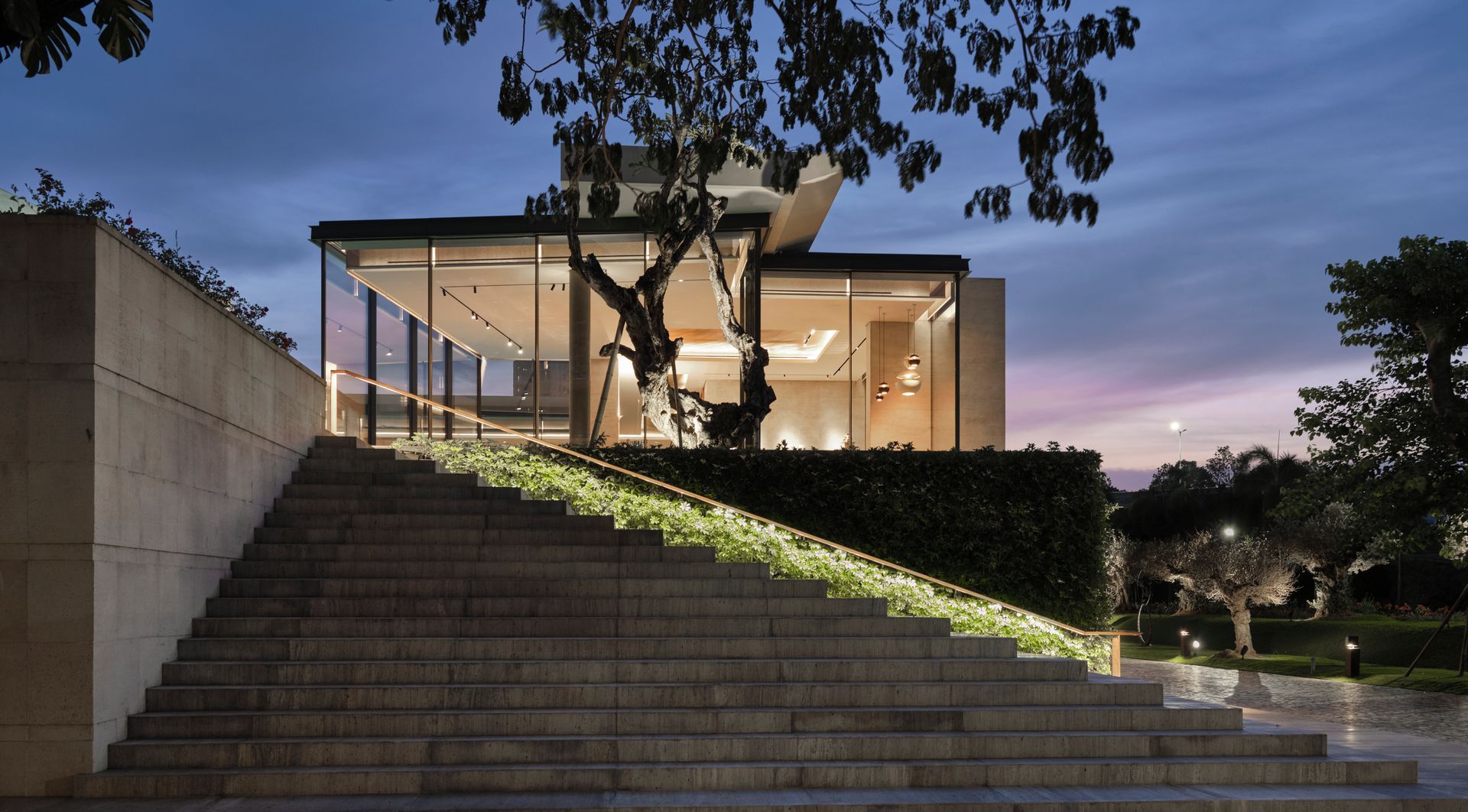 A modern home with OTIIMA large glass windows, viewed from the bottom of a grand staircase adorned with greenery and illuminated handrails. The evening sky adds a serene backdrop, enhancing the contemporary architectural design.