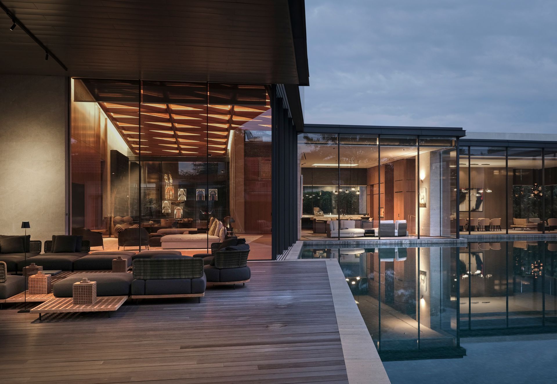 A sleek modern residence with OTIIMA expansive glass windows reflecting in a pool at dusk. The contemporary design is accentuated by warm interior lighting, offering a cozy and sophisticated view.