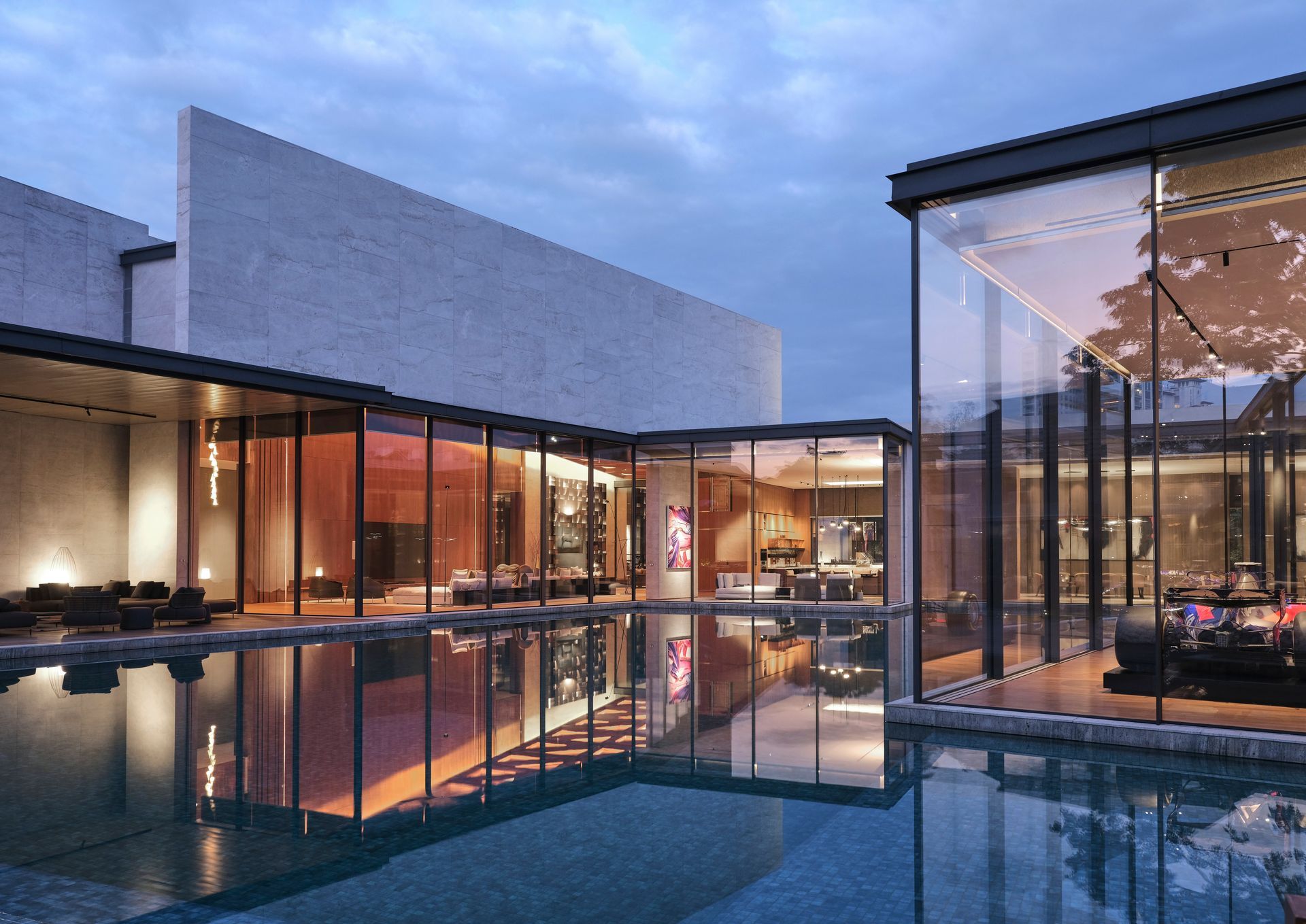 A sleek modern residence with OTIIMA expansive glass windows reflecting in a pool at dusk. The contemporary design is accentuated by warm interior lighting, offering a cozy and sophisticated view.