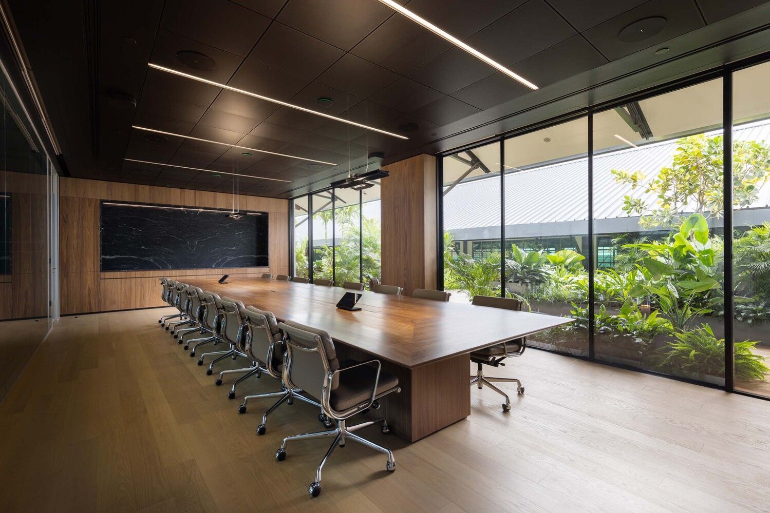 Modern meeting room with a long wooden table, ergonomic chairs, and large minimalist windows by OTIIMA offering views of a well-maintained garden.