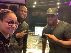 Three smiling people pose in a recording studio beside a mixing console and monitor.