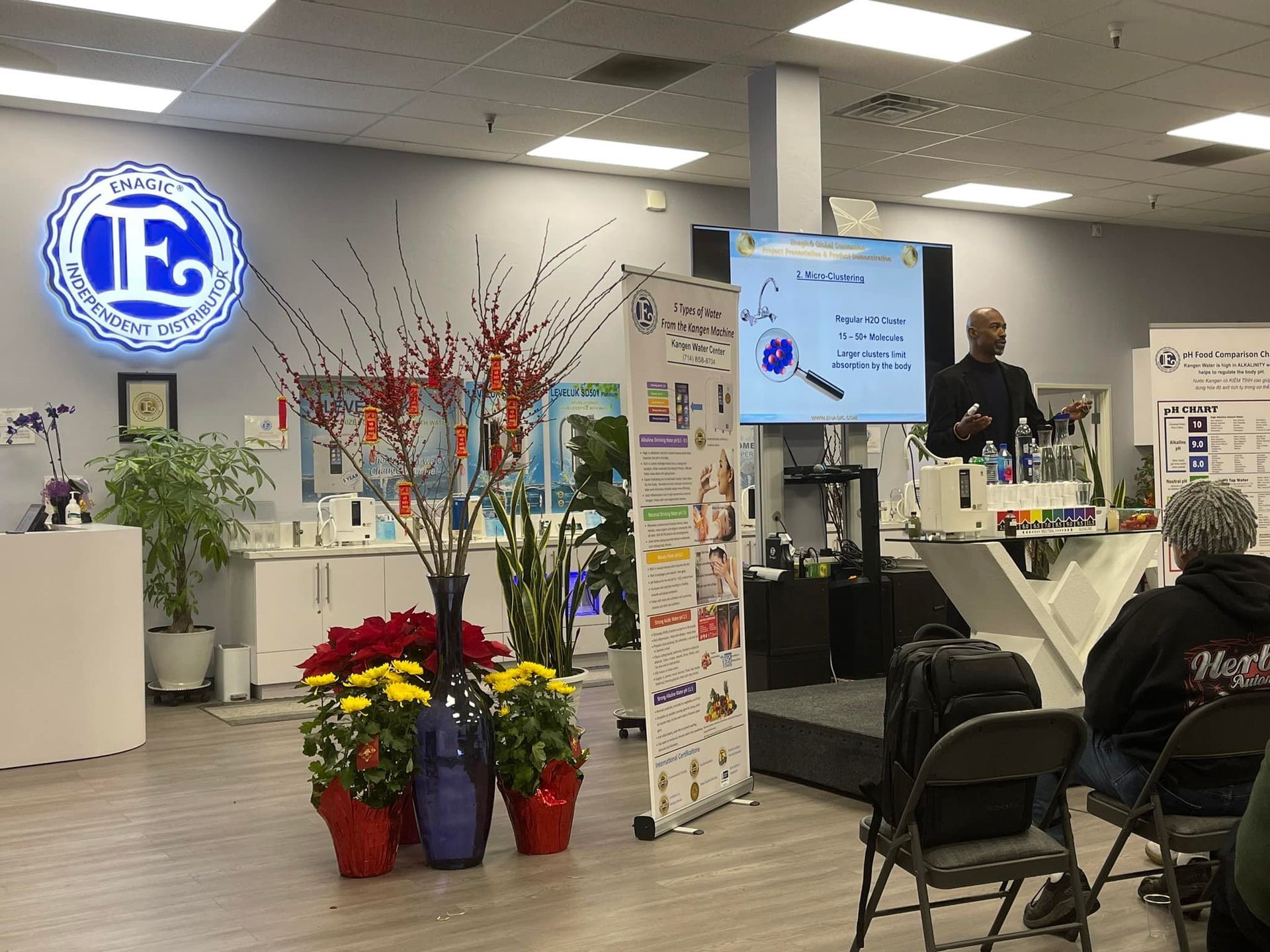 Man giving presentation in a retail space with display screens, products, and audience seated.