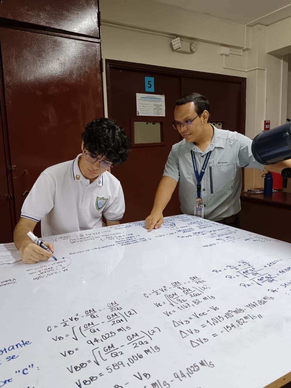 Dos hombres están sentados en una mesa escribiendo en una pizarra blanca.