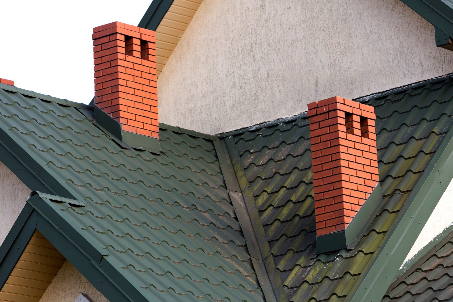 Roof with green shingles and two red brick chimneys against a light stucco wall