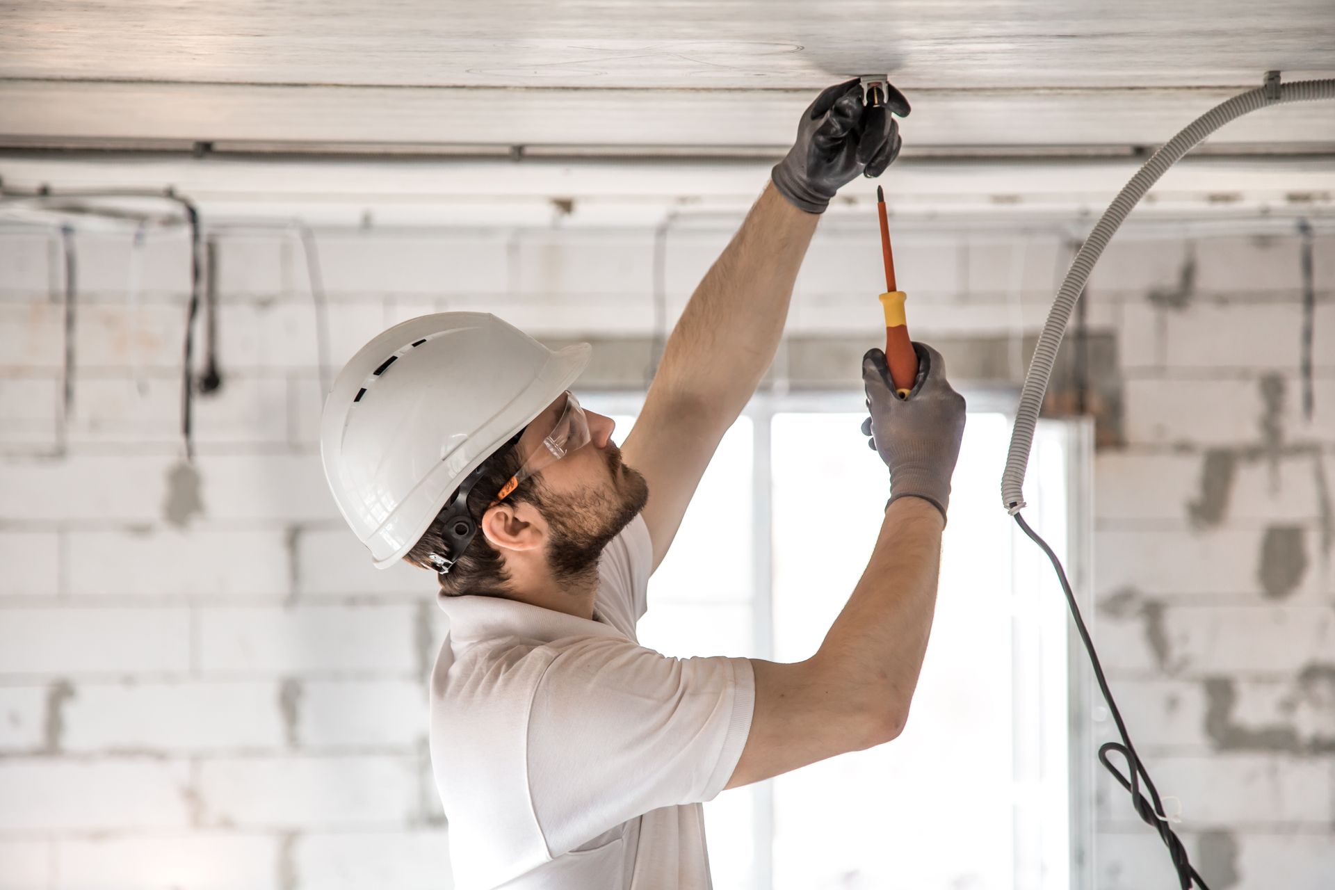 Worker in hard hat inspecting overhead wiring in a room with white brick walls