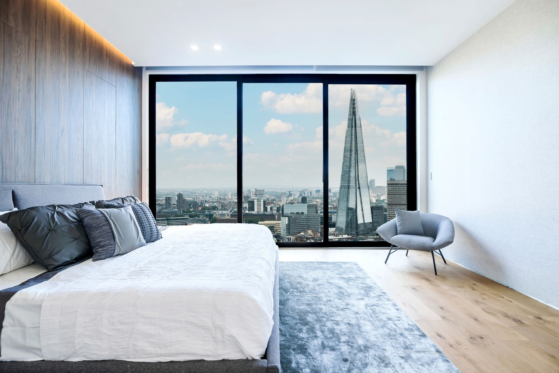 Modern bedroom with a large bed, blue rug, and floor-to-ceiling windows overlooking a city skyline