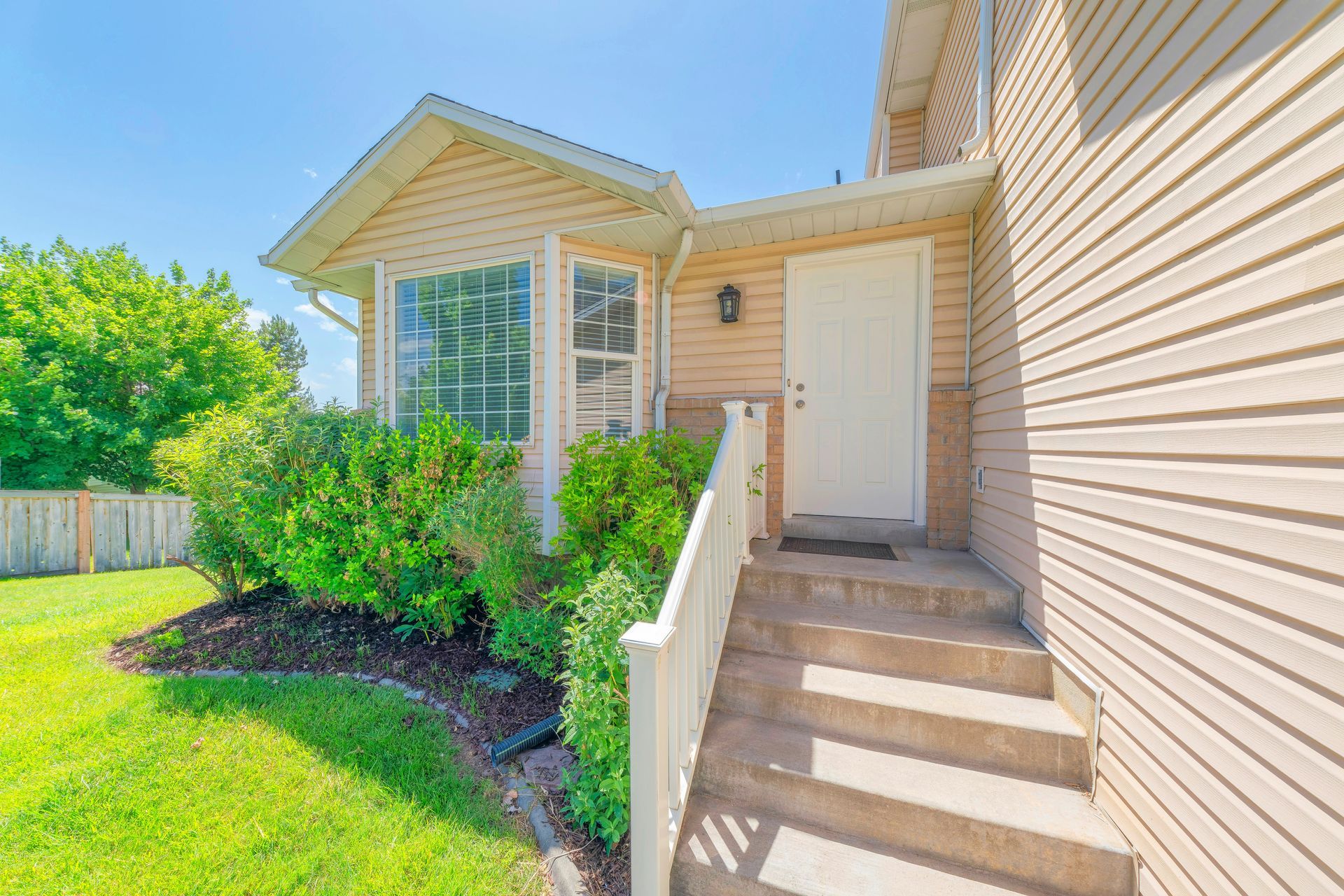 Sunlit townhouse entry with white stairs, beige siding, and green shrubs beside a small lawn