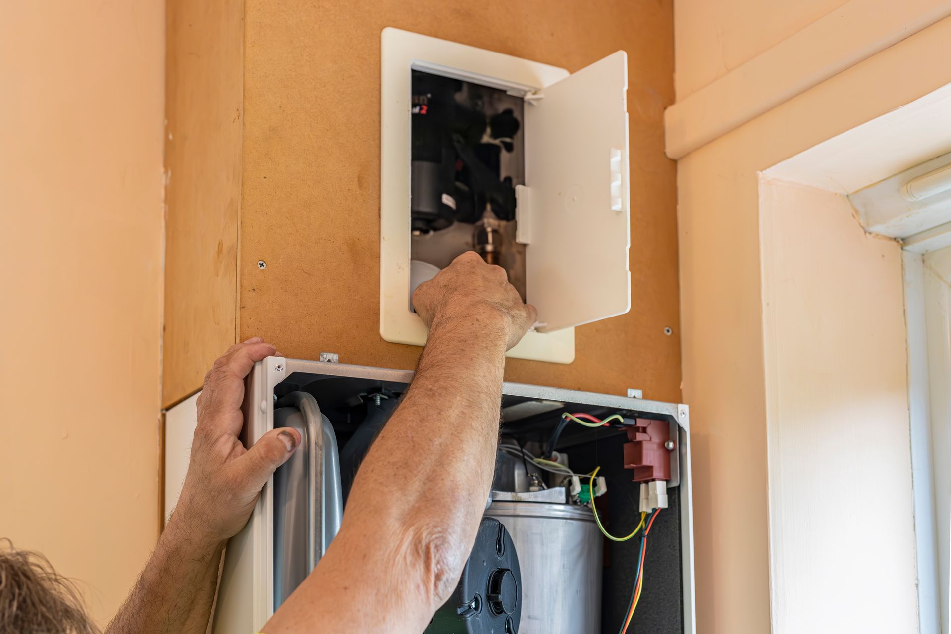 Electrician wiring an open wall panel near a ceiling access box in a peach-colored room