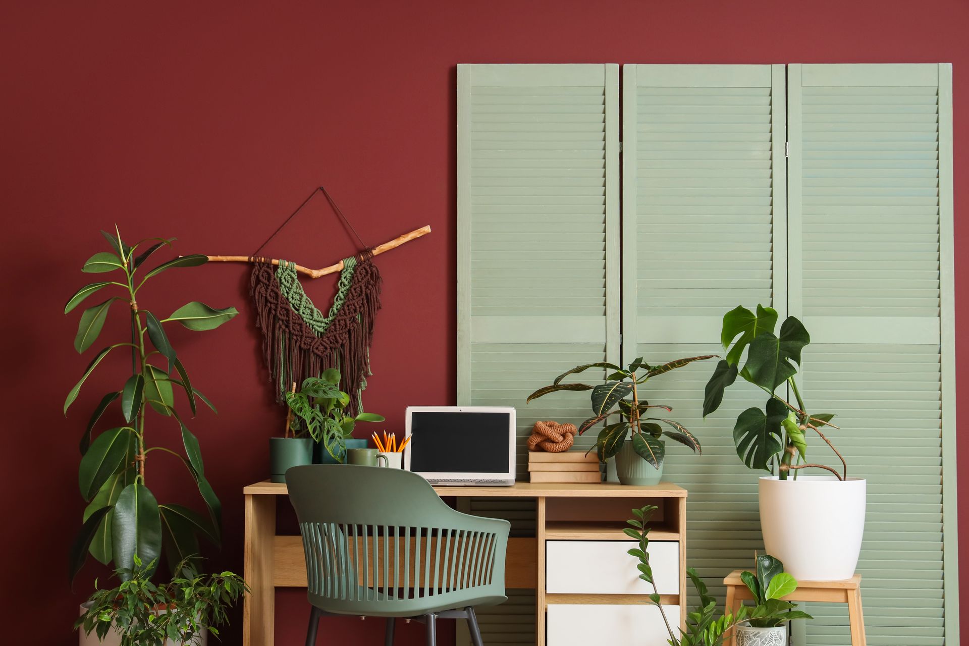 Cozy home office with laptop, chair, desk, and several potted plants against a red wall.