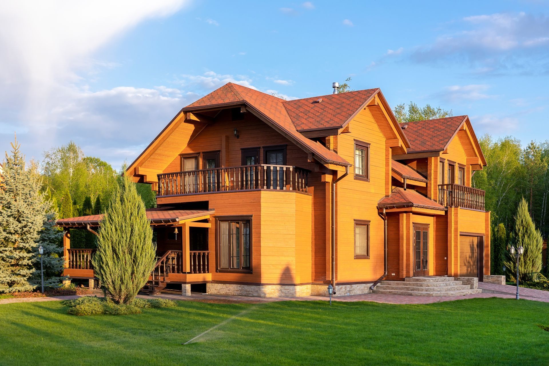 Two-story yellow house with red roof and balcony, surrounded by green lawn and trees under a blue sky