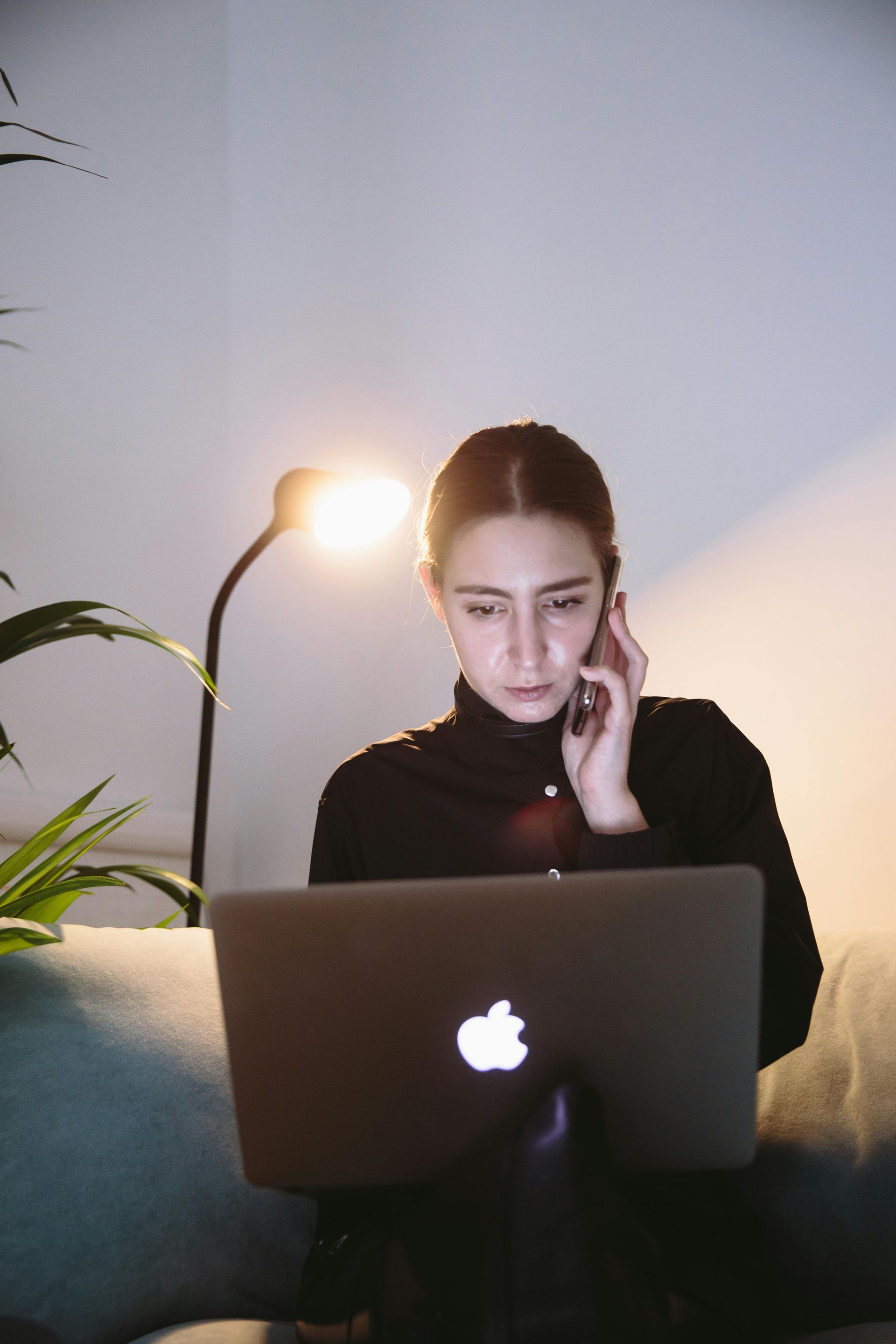 A woman is sitting on a couch talking on a cell phone while using a laptop computer.