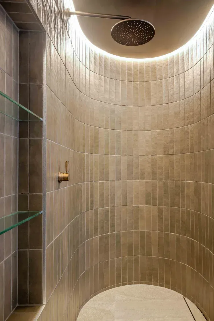 Curved beige tiled shower with a circular overhead light and glass shelf on the left