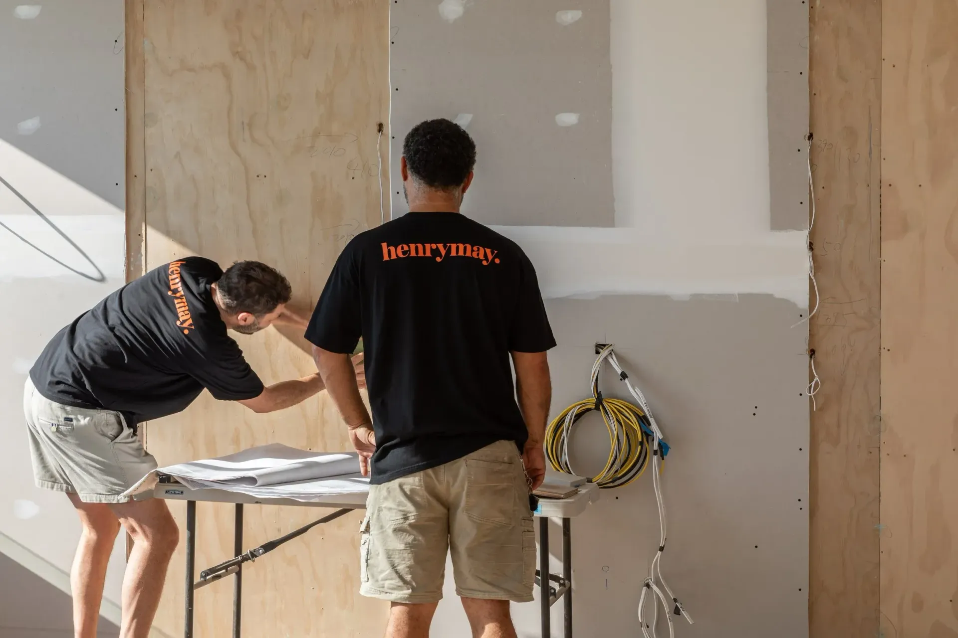 Two workers measuring drywall in an unfinished room, with tools and exposed wiring nearby.