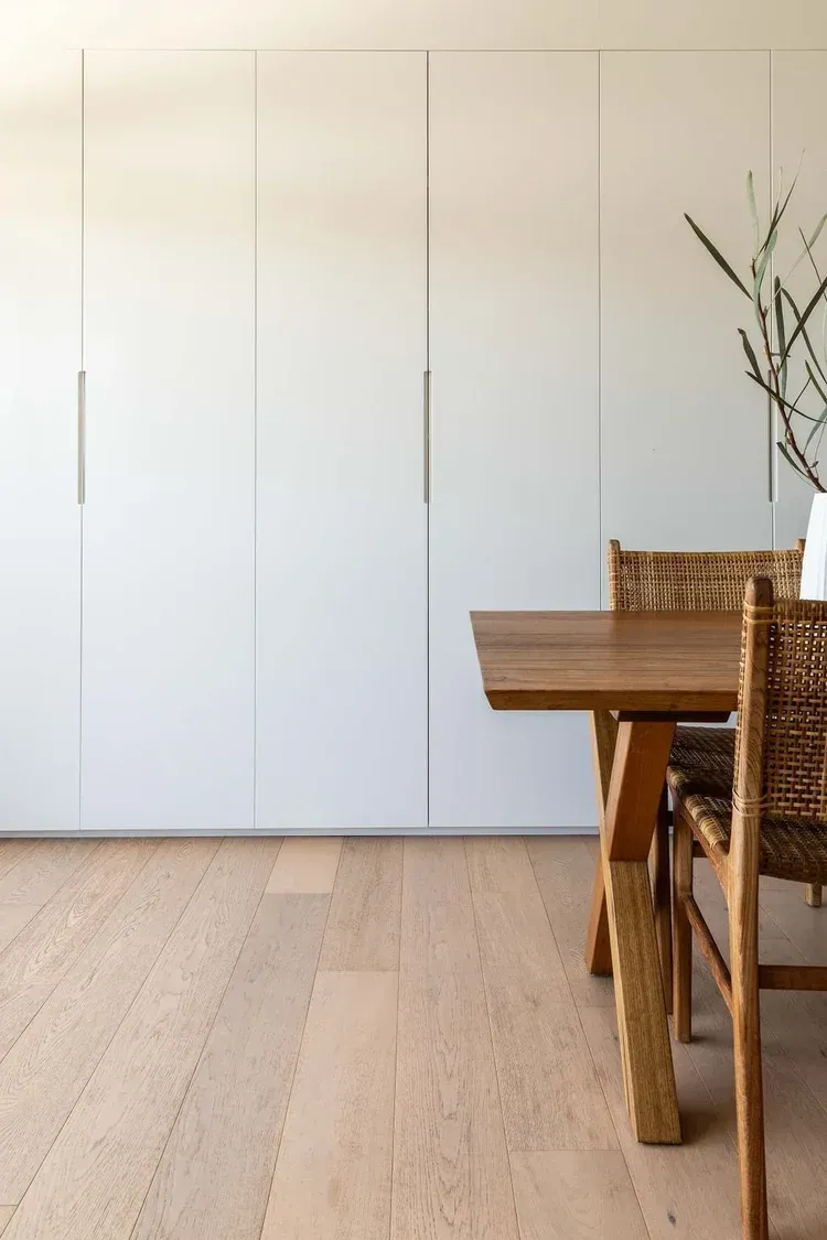 Minimal dining room with white closet doors, wooden table, wicker chairs, and a vase of branches