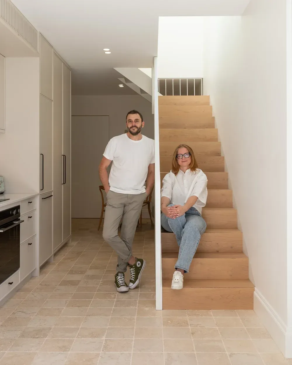 Two people posing in a bright hallway beside a staircase, with built-in cabinets and tiled floor.