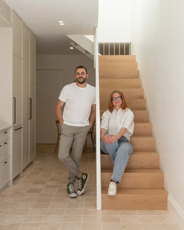 Two people posing in a bright hallway beside a staircase, one standing and one sitting on the steps.