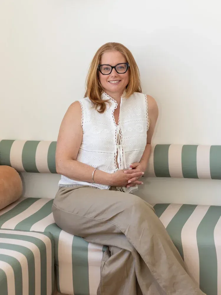 Woman sitting on a striped couch, smiling, wearing glasses and a white sleeveless blouse.