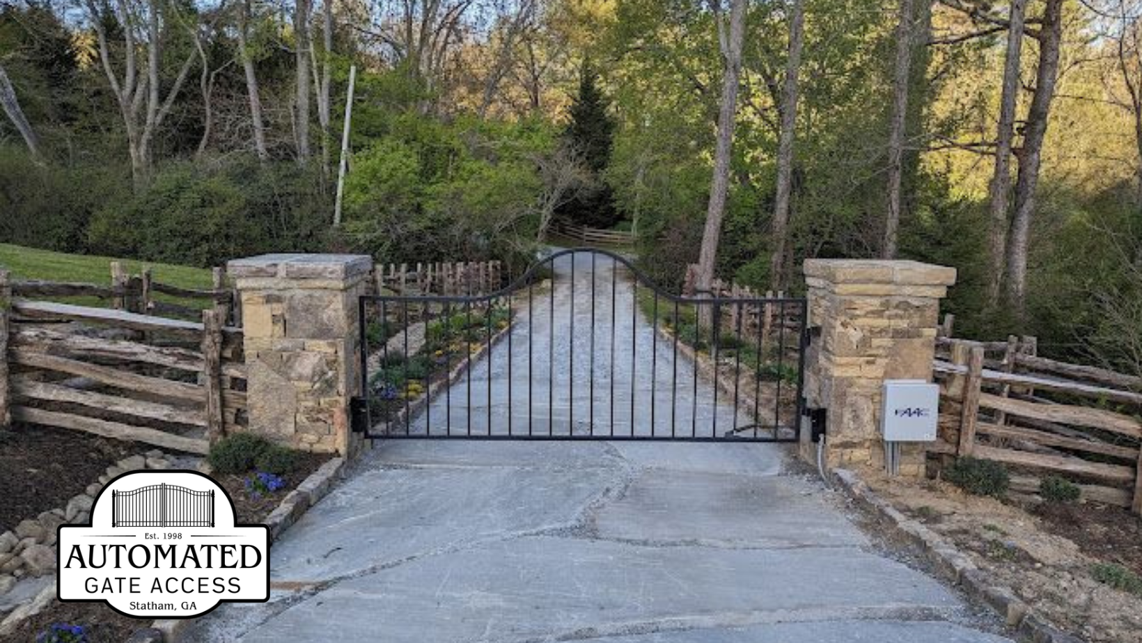 Black iron gate entrance to a driveway flanked by stone pillars, wooden fences, and trees.