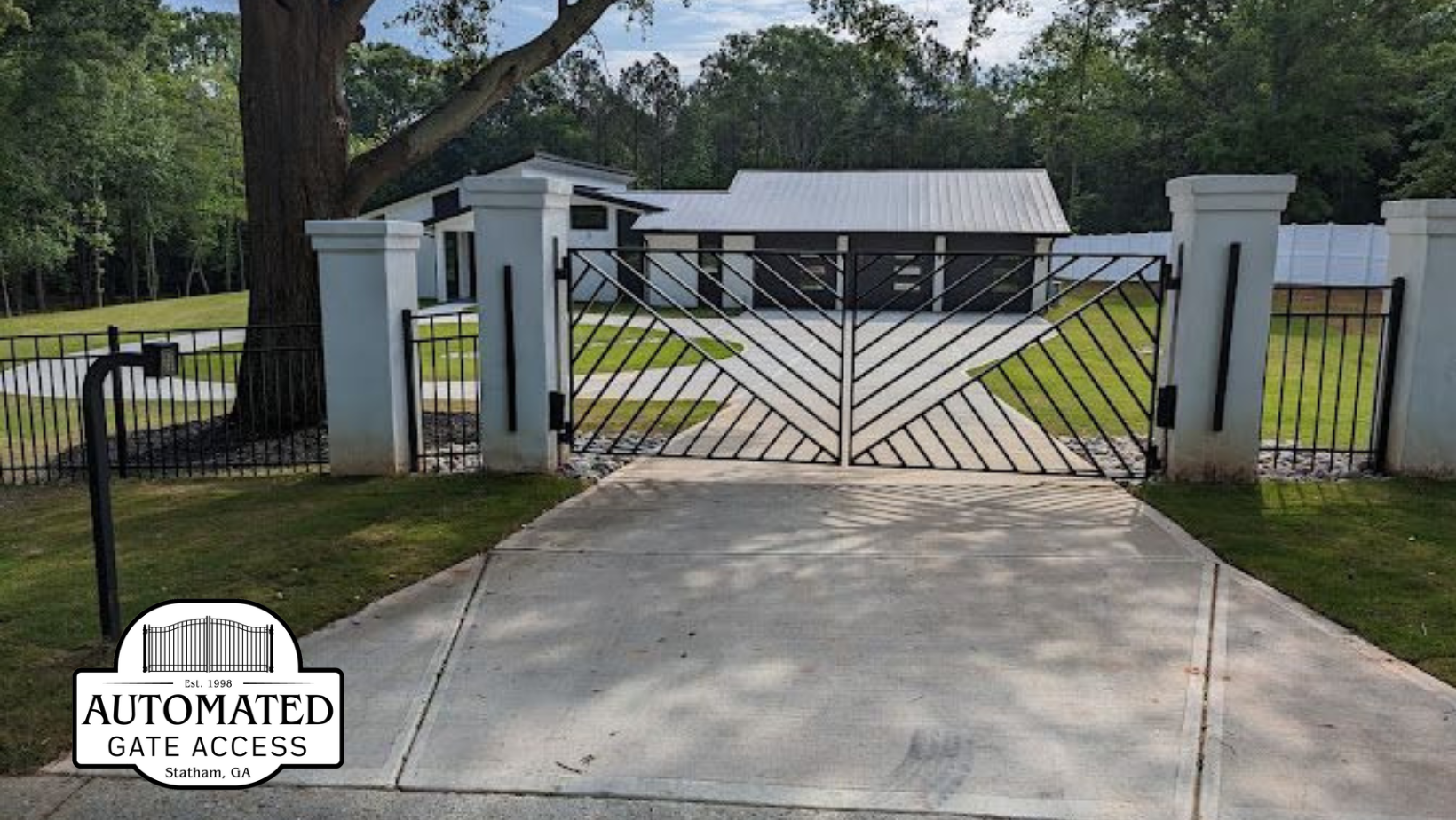 Driveway with decorative metal gate leading to a house, flanked by white pillars and black fencing.