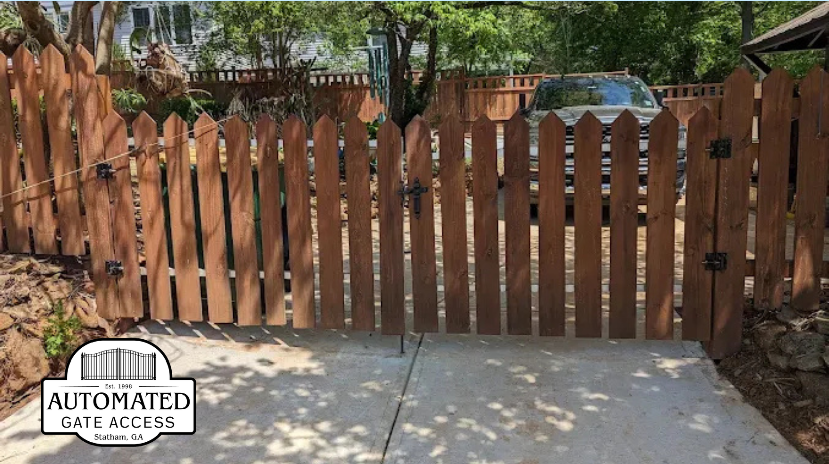 Wooden picket fence with open gate leading to a driveway with a parked car.