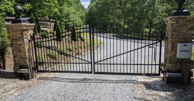 Black metal driveway gates between stone pillars; gravel driveway leads into trees.