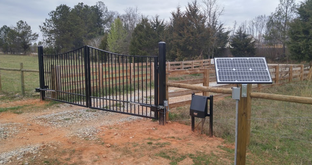 Black metal gate with solar panel for automated access, rural setting.