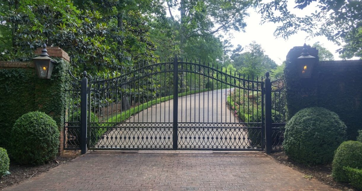 Black iron gate, opening to a driveway lined with hedges and trees.