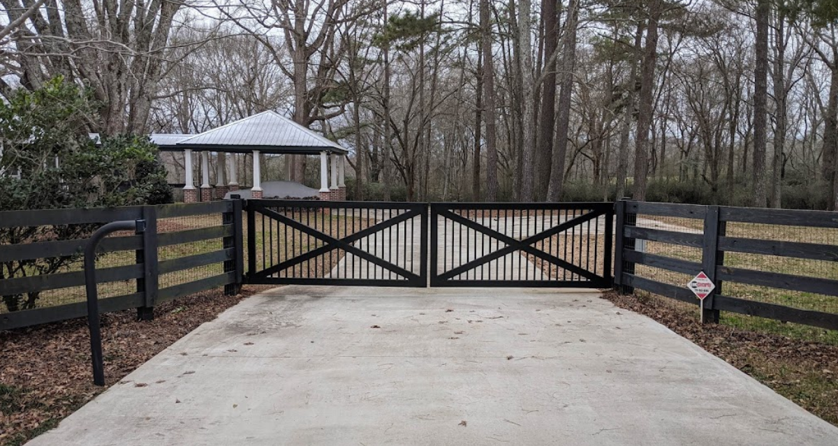 Black gate with diagonal supports, driveway, and wooden fence leading to a gazebo in a wooded area.