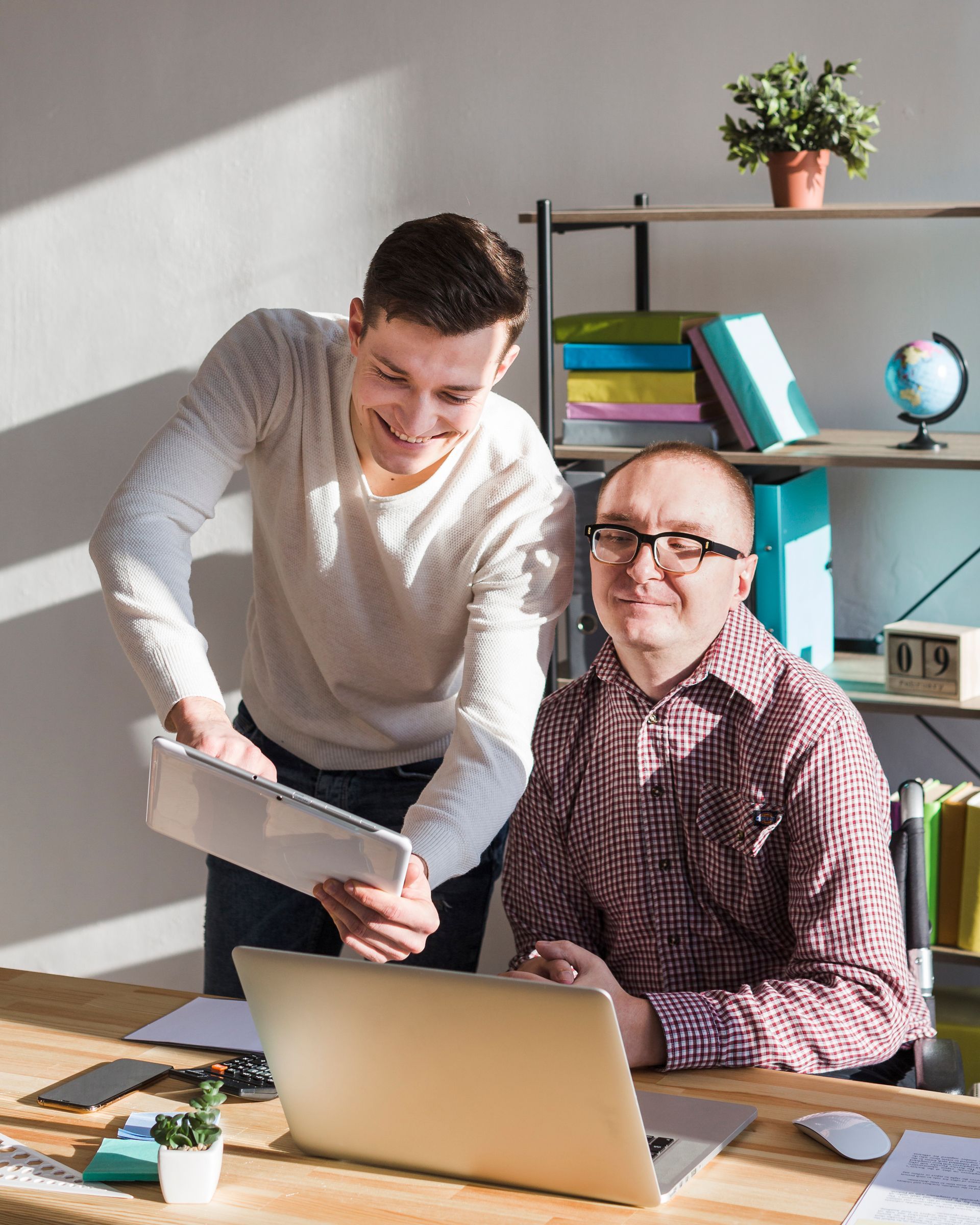 Two men are sitting at a desk looking at a laptop computer.
