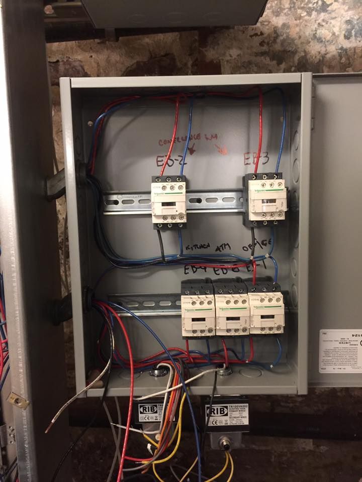 Control panel with wiring, contactors, and labeling against a brick wall.