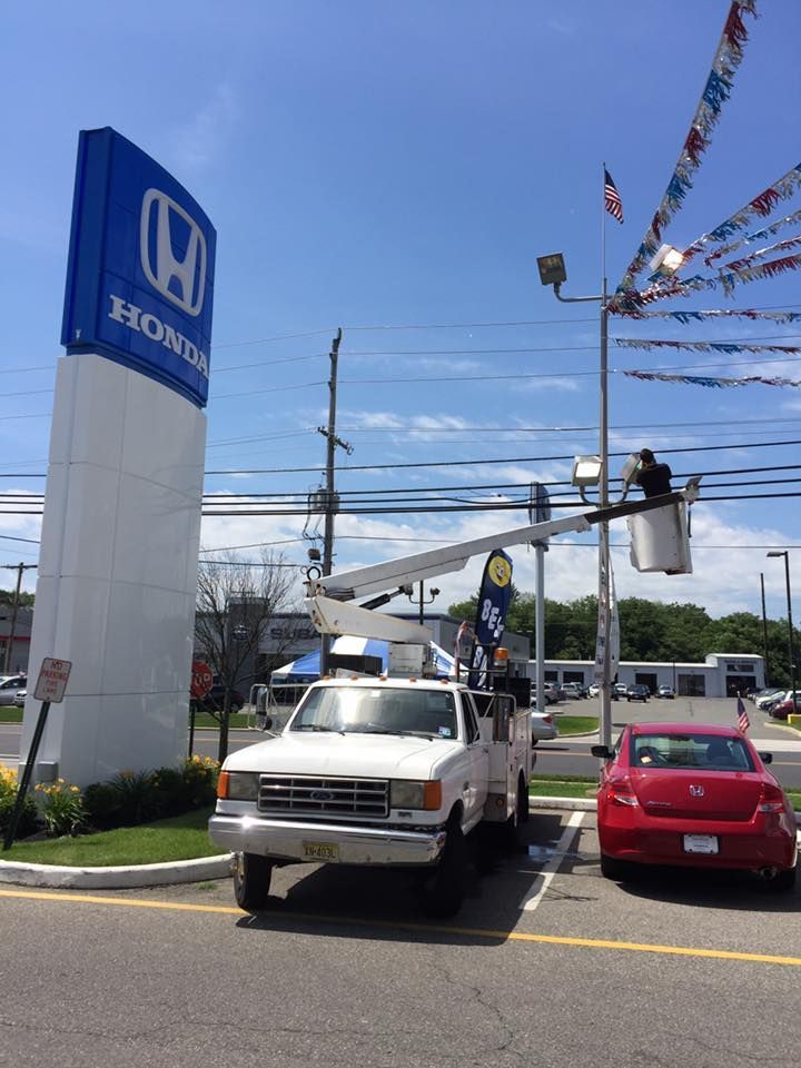 A white utility truck with its lift extended up to a light pole. A person is in the lift. A red car is parked next to it.