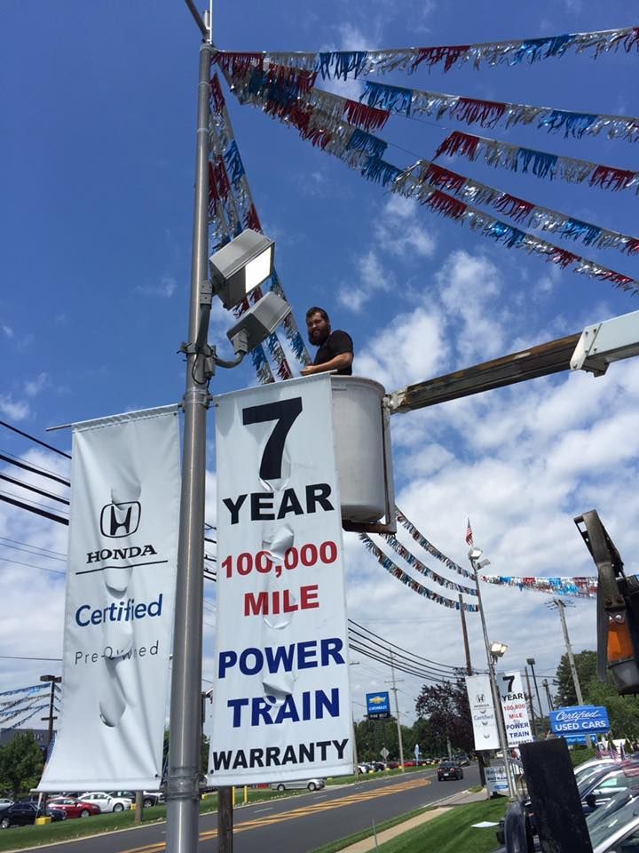 A person in a bucket lift fixing lights on a car dealership sign with red, white, and blue flags; sunny sky.