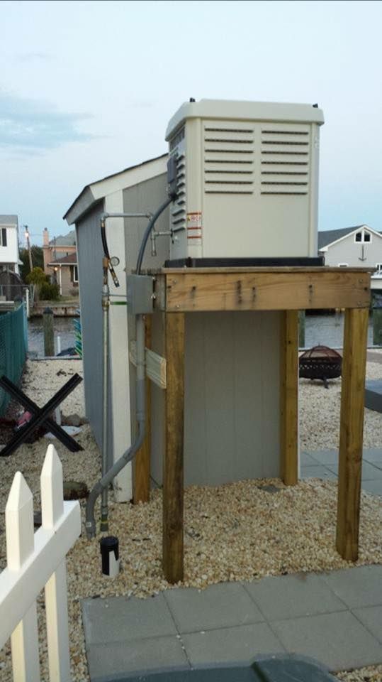 A standby generator installed on a wooden platform next to a shed in an outdoor setting.