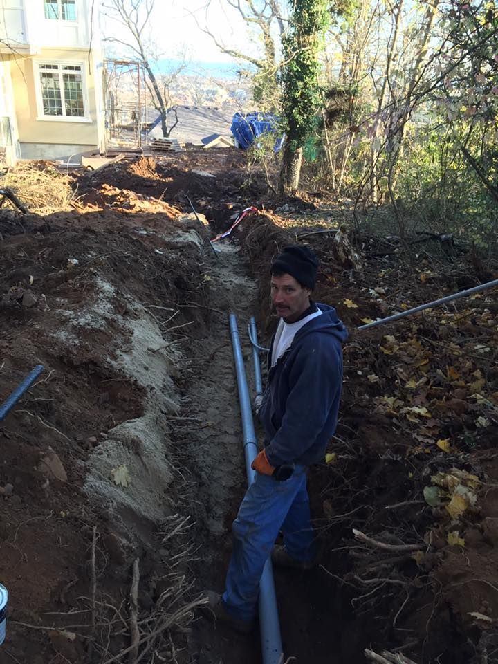 Man in a trench installing pipe. Dirt surrounds, blue sky and trees in background.