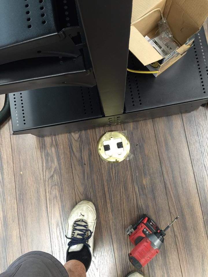 A person standing near a black rack, wooden floor, and a floor electrical outlet. A drill is visible.