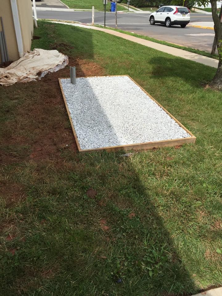 Gravel-filled rectangular area with wooden border and pipe. Adjacent to grass, sidewalk, and street with a car.