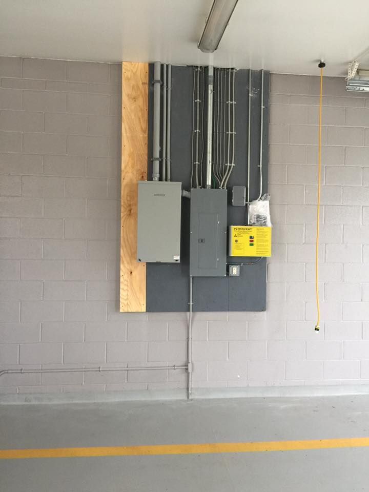 Electrical panel and conduit on gray wall. Yellow and gray boxes on plywood, gray floor.