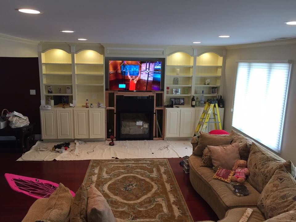 Living room with built-in cabinets around a TV, construction in progress. Tan couch, patterned rug, and a window with blinds.