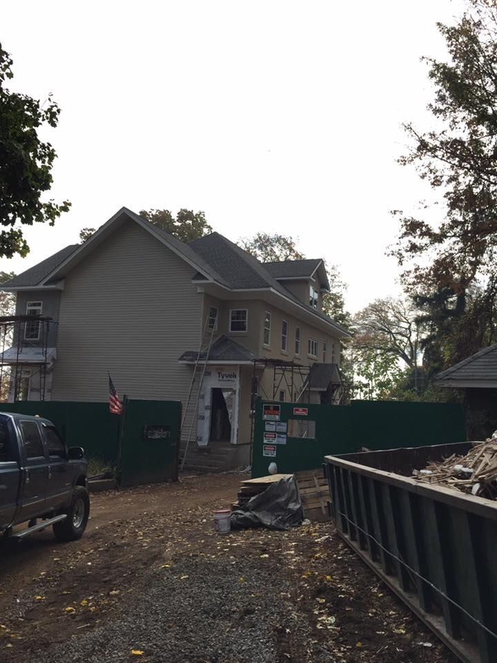 Two-story house under construction behind a green construction fence; a pickup truck and dumpster are in the foreground.