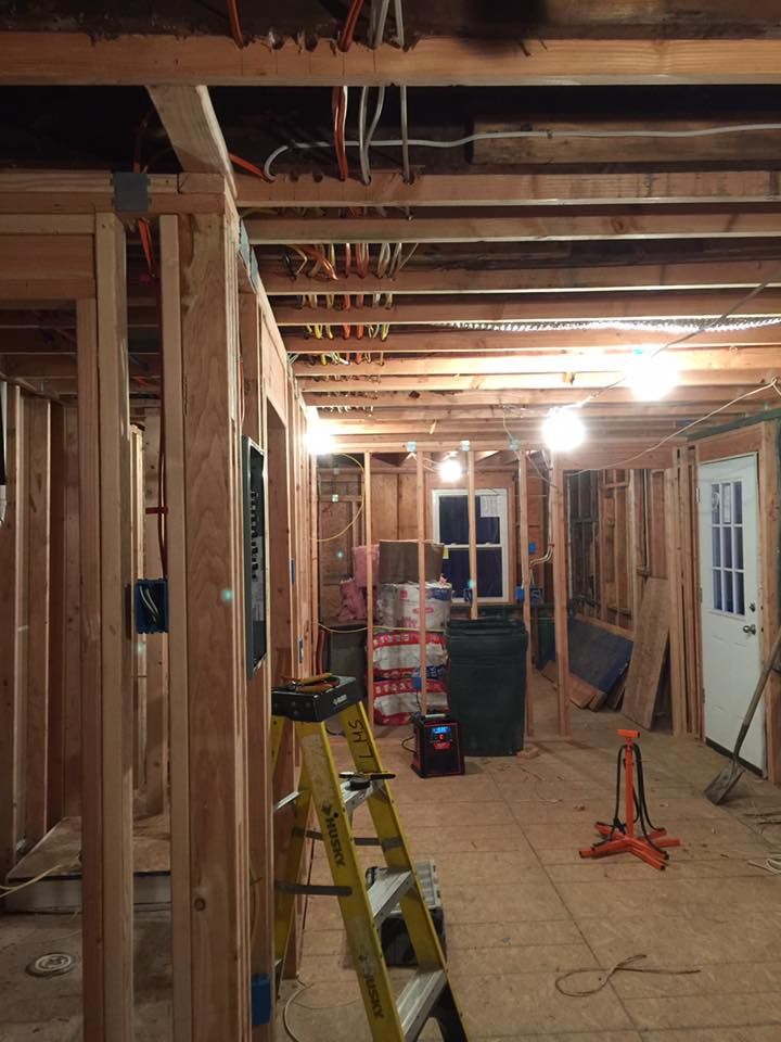 Interior view of a room under construction, with exposed wooden framing and electrical wiring. A ladder and tools are present.