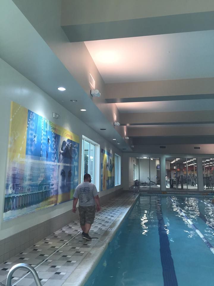 A man walks beside a pool in a gym, past large colorful wall art and windows with a view to exercise equipment.
