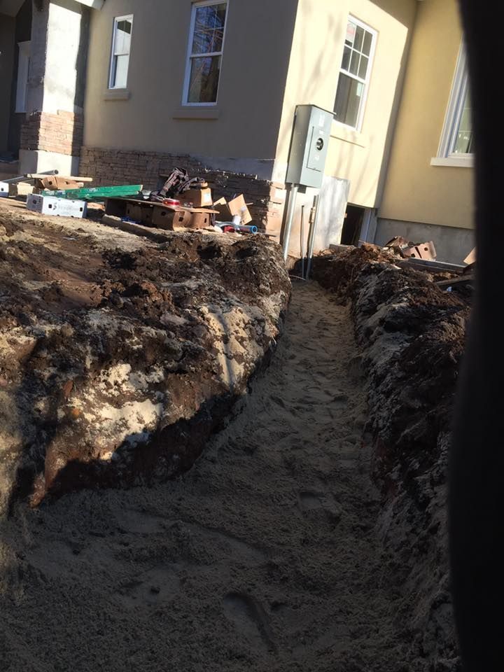 Trench dug beside a house, filled with sand. Electrical box visible on the wall. Construction site.